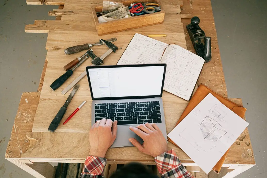 Hands typing on a laptop at a workbench beside tools and sketches